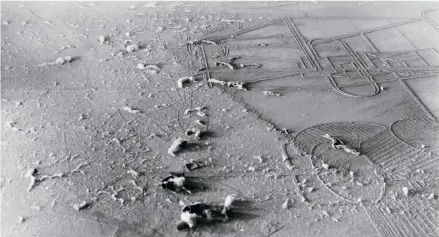 Fotografía aérea en blanco y negro de un paisaje árido con marcas y surcos que forman figuras geométricas, posiblemente una obra de land art o intervención en el terreno.