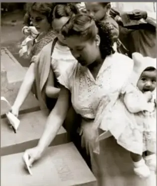 Fotografía en sepia de 1955: mujeres haciendo sufragio frente a una urna, una de ellas con una niña en brazos. Crédito: Archivo Gráfico de El Nacional, Acervo INEHRM.
