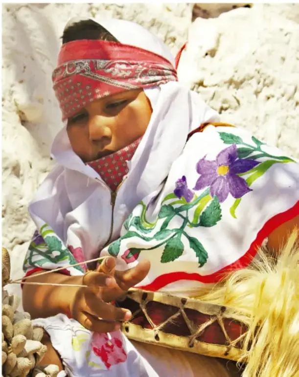 Fotografía de una niña rarámuri con pañuelo rojo en la cabeza y vestido blanco bordado con flores moradas y rojas; trabaja con hilos o fiber y sostiene una pequeña tambora o tambor de aro, junto a sogas de fibra.