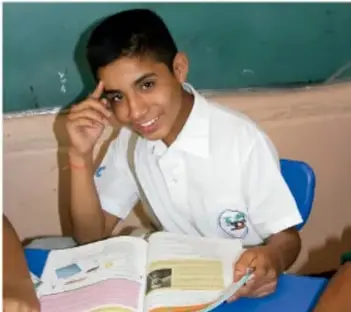 Fotografía de un estudiante de secundaria con uniforme blanco, sentado en su escritorio, pensativo con la mano en la frente, frente a un libro abierto; pizarra verde al fondo.