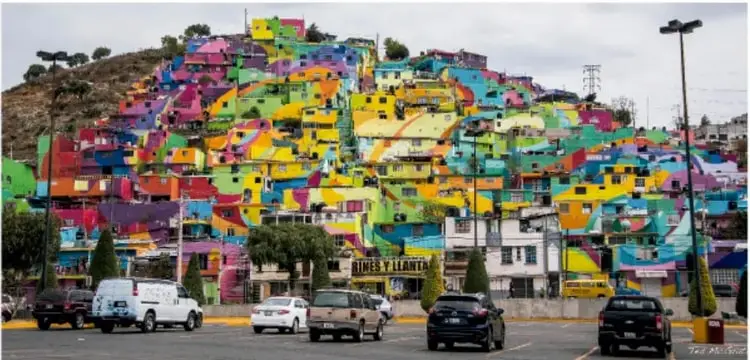 Fotografía panorámica de Las Palmitas, Pachuca, Hidalgo: ladera cubierta de casas pintadas de colores vivos (mural urbano del Germ Collective), estacionamiento con automóviles al frente, fotografía de Ted McGrath.