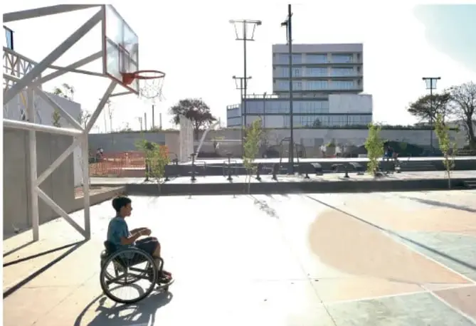 Fotografía de una persona en silla de ruedas en una cancha de básquetbol al aire libre, con canasta, élite y edificios modernos al fondo; representa la inclusión deportiva en el espacio comunitario.