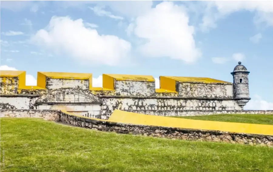 Fotografía a color de los restos de la muralla de Campeche: muro de piedra con almenas pintadas de amarillo, garita de vigilancia redonda y césped al frente, bajo un cielo con nubes.