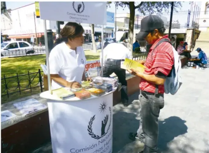 Fotografía de un módulo de la Comisión Nacional de los Derechos Humanos en la vía pública, con una funcionaria atendiendo a un ciudadano que revisa folletos informativos.