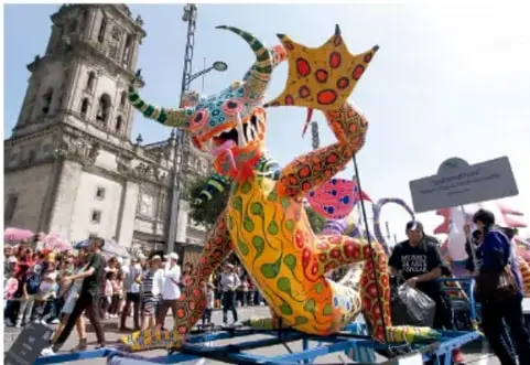 Fotografía de una calle con una gran figura artística colorida (alebrije o monstruo de papel estilo alebrije) en un carro alegórico, rodeada de personas, con un templo al fondo; representa una manifestación artística en espacio público.