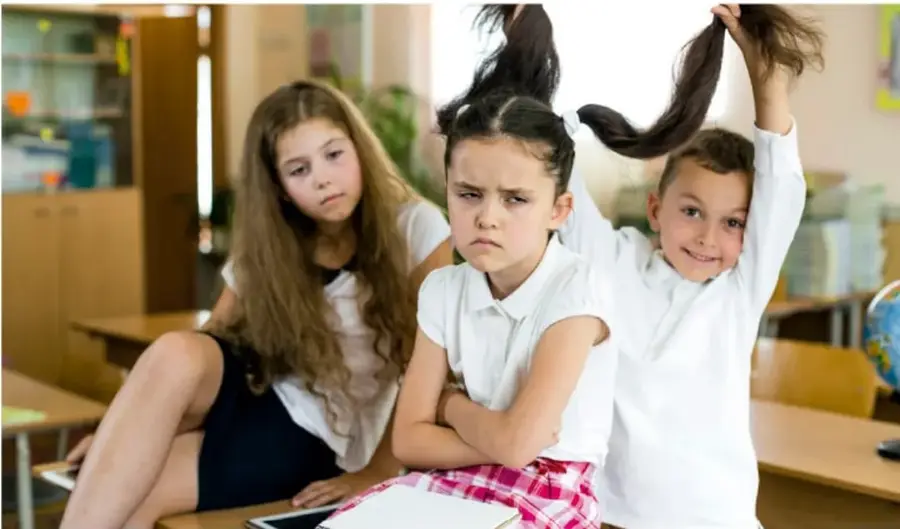 Fotografía de tres estudiantes en un salón de clases: una niña con expresión de enojo y brazos cruzados, un niño detrás jalándole el cabello con sonrisa, y otra niña observando con gesto de molestia. Representa situaciones escolares que provocan emociones.