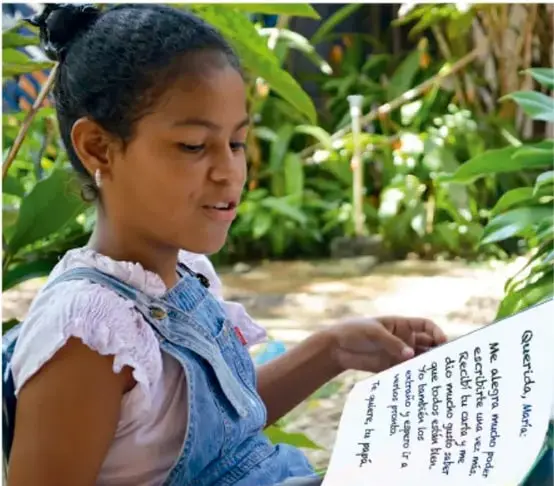 Fotografía de una niña de cabello recogido, con blusa blanca y overol de mezclilla, leyendo una carta al aire libre entre plantas.