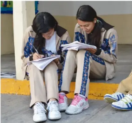 Dos estudiantes sentadas en el piso, con uniforme escolar, escribiendo en sus cuadernos; en el suelo se ven sus mochilas y zapatos de compañeros, ilustrando la redacción de acuerdos asamblearios.