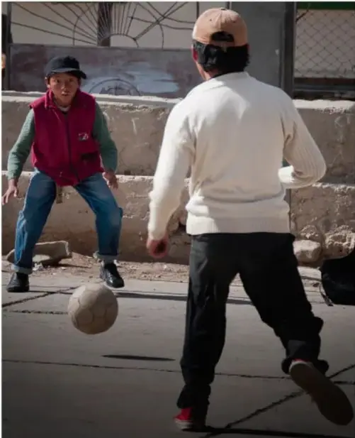 Dos niños juegan fútbol en un patio; uno patea el balón y otro espera como portero.