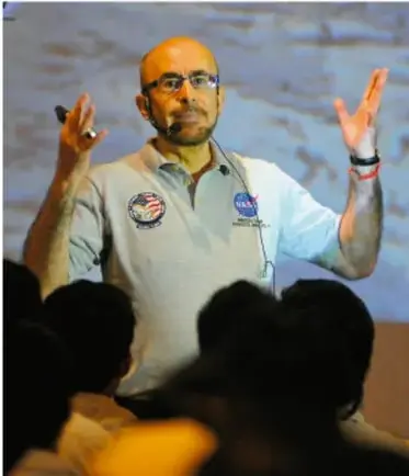 Fotografía de Rodolfo Neri Vela dando una conferencia, con camisa clara y logotipos de la NASA.
