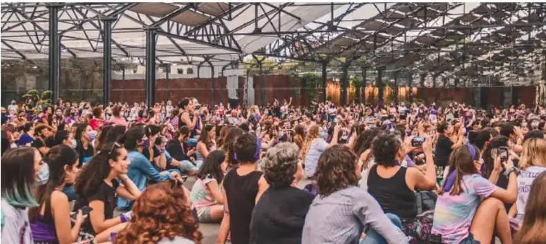 Fotografía de una asamblea o reunión comunitaria multitudinaria bajo una estructura techada, con muchas personas sentadas y algunas de pie grabando con teléfonos móviles, representando la participación ciudadana.