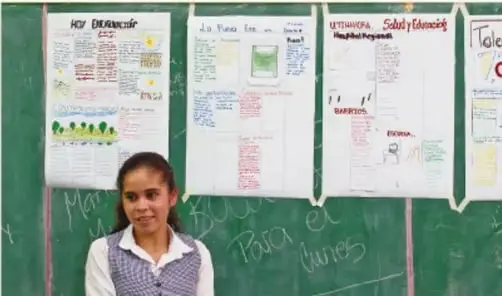 Fotografía de una estudiante frente a una pared verde con carteles escolares escritos a mano sobre diversidad cultural y lenguas.