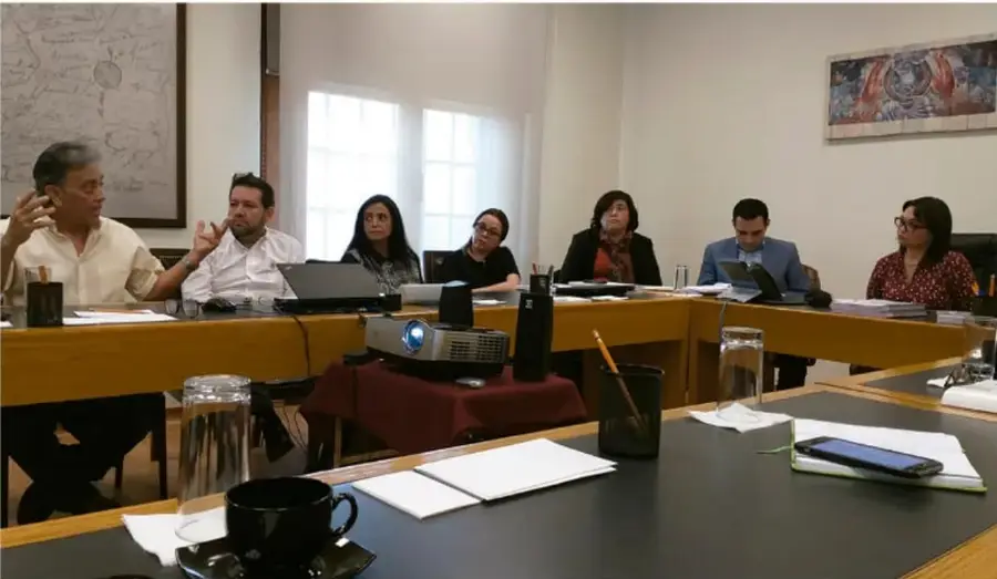 Fotografía de una reunión de trabajo en un salón con una mesa larga, proyector y varias personas sentadas participando en una evaluación o junta de mejora continua.