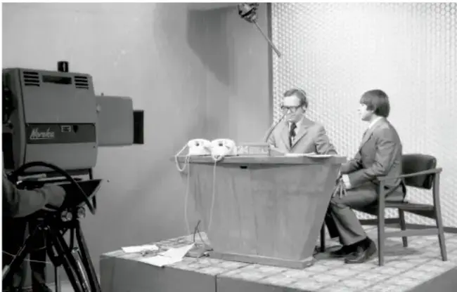 Fotografía en blanco y negro de un set de televisión con dos hombres sentados en un escritorio, uno frente a una cámara.