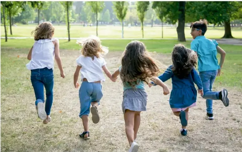 Fotografía de cinco niños corriendo y tomados de la mano en un parque con árboles y césped, como ejemplo de actividad física y convivencia comunitaria.