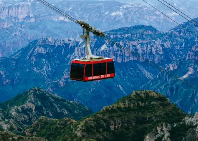 Cabina roja de teleférico colgada de cables que cruza el cielo azul sobre montañas verdes escarpadas de la barranca.