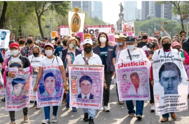 Fotografía de una marcha ciudadana en la Ciudad de México: manifestantes con cubrebocas portan retratos de los estudiantes desaparecidos de Ayotzinapa y carteles con las consignas '¡Justicia!', '¡Vivo se lo llevaron, vivo lo queremos!' y 'Ayotzinapa se lo llevaron'. Al fondo se observan edificios y árboles de la avenida.