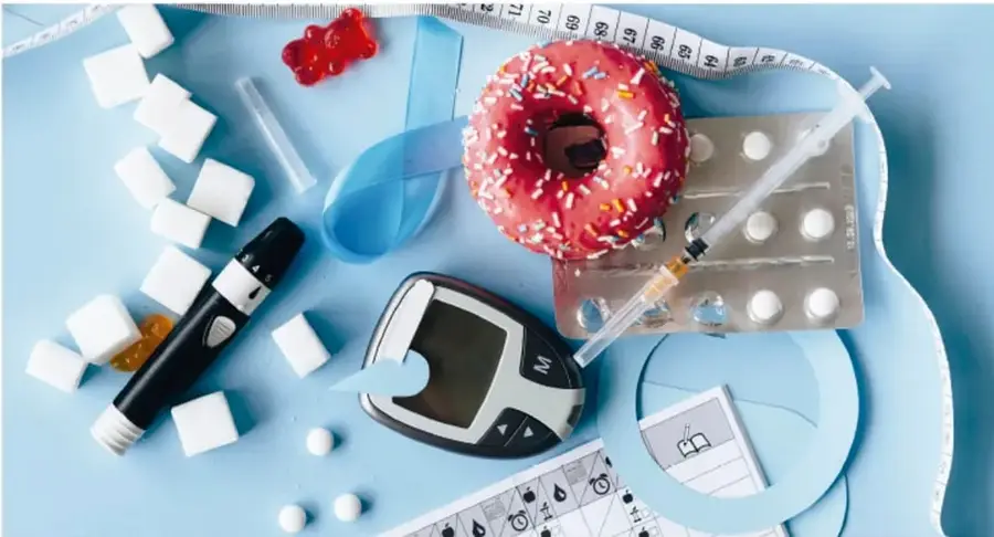 Composición fotográfica sobre fondo azul con una dona con sprinkles, cubos de azúcar, un glucómetro con tiras reactivas, una jeringa, blíster de pastillas, un blister de inyección, gominolas de oso, cinta métrica y una agenda de control de glucosa: elementos que representan la diabetes.