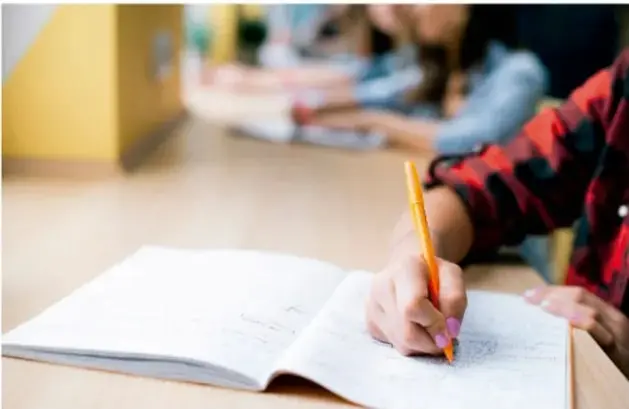 Fotografía de un(a) estudiante escribiendo con lápiz amarillo en un cuaderno abierto sobre un escritorio, con otros estudiantes difuminados al fondo, en un aula.