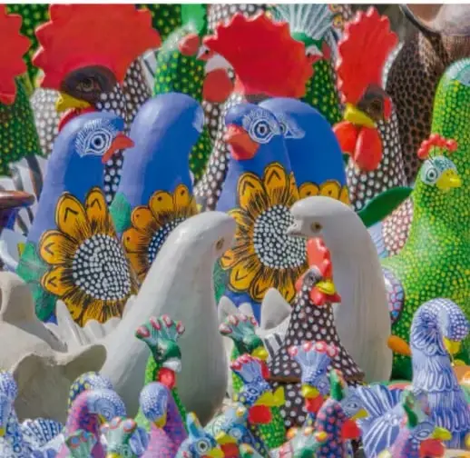 Fotografía de artesanías mexicanas de colores vibrantes: figuras de cerámica y talla en madera (gallos, palomas) decoradas con flores y patrones multicolor, ilustrando las manifestaciones artísticas del país.
