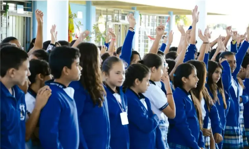 Fotografía de un grupo de estudiantes de secundaria con uniforme azul levantando las manos, en actitud participativa dentro de un espacio escolar.