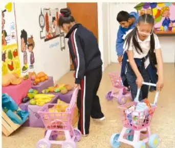 Fotografía de estudiantes en un mercado escolar manipulando frutas y verduras en carritos de juguete.