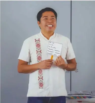 Fotografía de un estudiante de telesecundaria sonriendo y mostrando una libreta con anotaciones, con camisa blanca bordada.