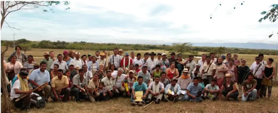Fotografía de un grupo numeroso de personas, aparentemente integrantes de una comunidad indígena, reunidas al aire libre en un campo. Visten ropas tradicionales y sostienen algunos objetos. Al fondo se observan árboles y un paisaje rural.