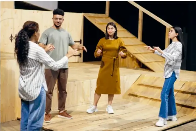 Fotografía de cuatro estudiantes sobre un escenario de madera representando una escena teatral/audiovisual, con la leyenda: 'La cantidad de personajes involucrados da cuenta de que no es fácil hacer audiovisuales; sin embargo, comprender los pasos y lo que hace cada participante es necesario para llevarlo a cabo en el aula.'