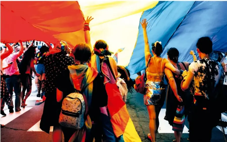 Grupo de personas, vistas de espaldas, marchan por una calle sosteniendo una gran bandera con franjas de colores vivos (rojo, amarillo, azul) y elementos del arcoíris; llevan mochilas y ropa colorida. La escena sugiere una marcha pacífica por derechos y diversidad.
