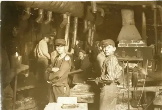 Fotografía en blanco y negro de trabajadores en una fábrica durante el siglo XIX. Se observan hombres y niños trabajando en un ambiente industrial con maquinaria y poca iluminación.