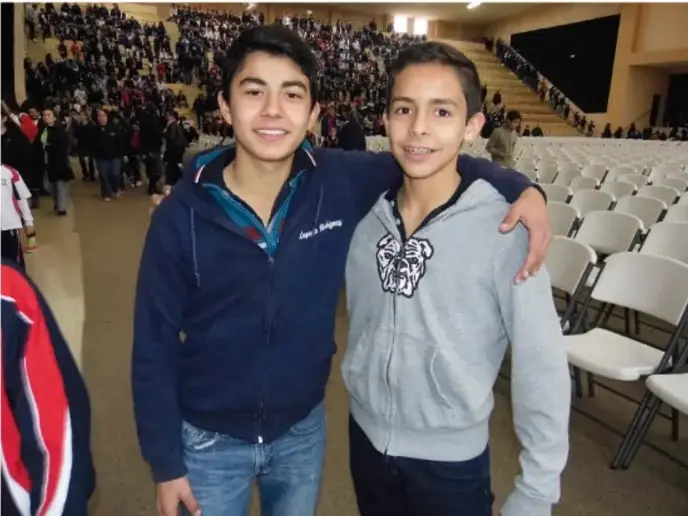 Fotografía de dos adolescentes sonriendo y abrazados en un auditorio con público, ilustrando la expresión de emociones positivas en un entorno comunitario escolar.