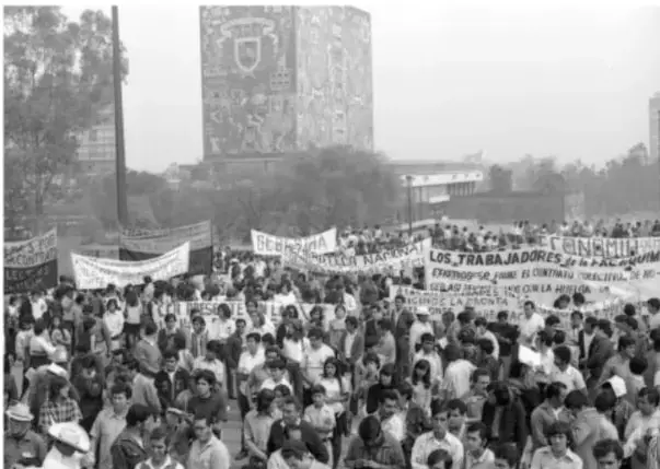 Fotografía en blanco y negro de una manifestación magisterial en la Ciudad de México, con una pancarta que dice 'LOS TRABAJADORES DE LA EDUCACIÓN...' frente a un edificio con un mural.