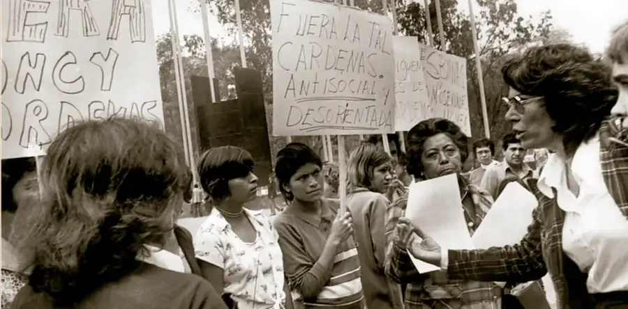 Fotografía en blanco y negro de una protesta de mujeres en México, 1975. Se ven mujeres con carteles; uno de ellos dice 'FUERA LA TAL CÁRDENAS, ANTISOCIAL Y DESORIENTADA' y otro menciona el 'AÑO INTERNACIONAL DE LA MUJER'. Una mujer con lentes conversa con otras que sostienen papeles. Pie de foto: protesta en contra de Nancy Cárdenas y de las conferencias del Año Internacional de la Mujer, 1975. Acervo INEHRM.