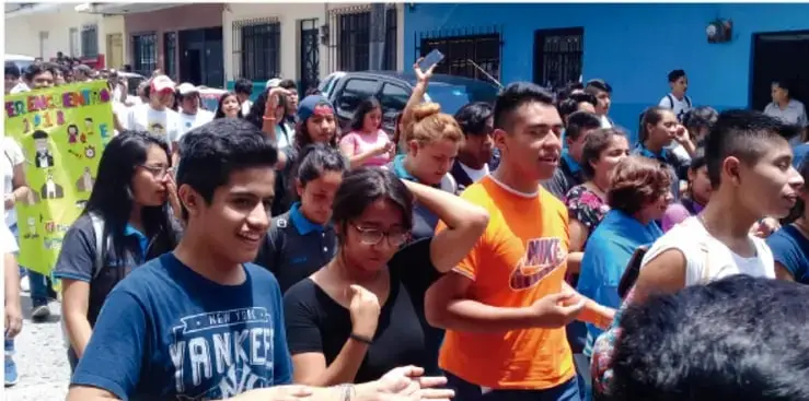 Fotografía de una marcha o manifestación juvenil en una calle. Se observa a un grupo numeroso de adolescentes y jóvenes caminando, algunos con pancartas y carteles. Destacan jóvenes con playeras de colores vivos, expresiones de entusiasmo y participación colectiva.