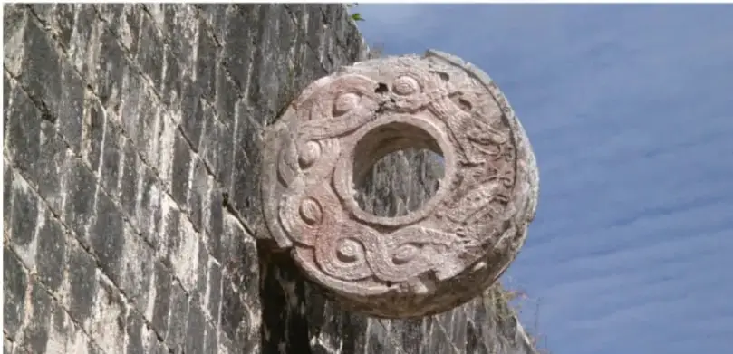 Fotografía de un anillo de piedra monumental incrustado en un muro, típico de las canchas del juego de pelota prehispánico, con fondo de agua/espacio azul.