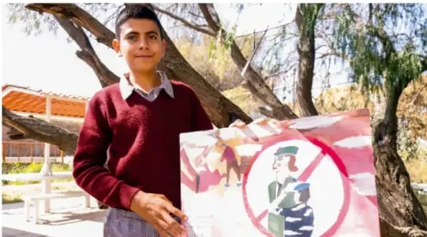 Fotografía de un estudiante de telesecundaria sosteniendo un cartel con un símbolo de prohibición sobre una imagen (mensaje de prevención), en un espacio escolar con árboles al fondo.