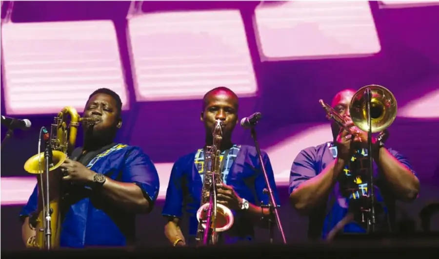Fotografía de tres músicos afrodescendientes tocando saxofón, trompeta y trombón sobre un escenario con iluminación morada; visten trajes azules con estampados afrocaribeños y hay micrófonos y atriles con partituras.