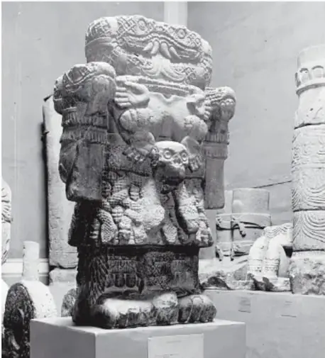 Fotografía en blanco y negro de la escultura original de Coatlicue exhibida en un museo, sobre pedestal, con sus serpientes, collar de corazones y manos tallados en piedra.