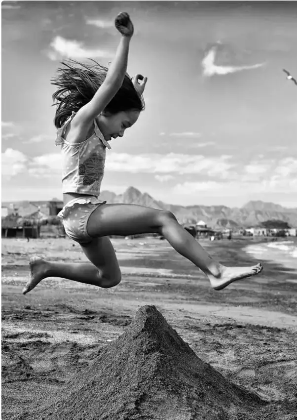 Fotografía en blanco y negro de una niña saltando en la playa: brazos levantados, rodillas flexionadas y cuerpo en el aire sobre un montículo de arena.