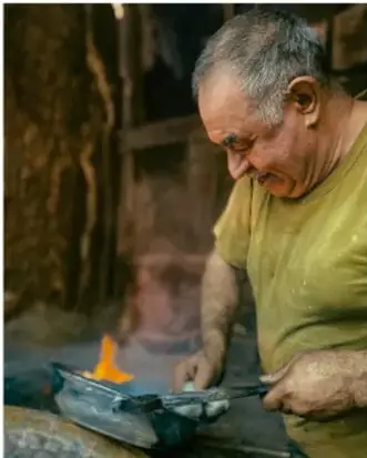Fotografía de un artesano mayor trabajando con metal y fuego en un taller, ilustrando la práctica artística comunitaria.