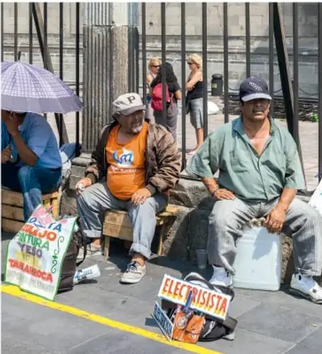 Fotografía de personas sentadas en una banqueta con carteles que dicen 'ELECRICISTA' y otros letreros, en un contexto urbano; parecen trabajadores informales o migrantes.