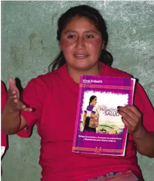 Fotografía de una mujer indígena sonriente con rebozo rojo que sostiene la publicación 'Kinal Antzetik: Guía para promotores de salud'.