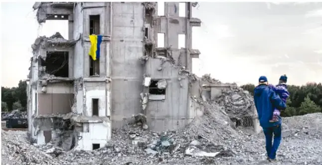 Fotografía de un edificio residencial destruido por bombardeos en Ucrania, con la bandera del país asomando por una ventana en ruinas; en primer plano, un adulto carga a una niña mientras caminan entre los escombros.