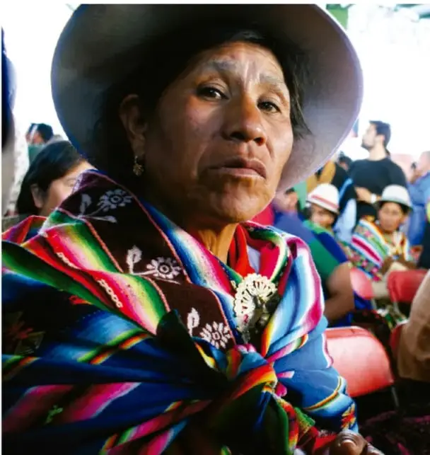 Fotografía de una mujer indígena de edad adulta, con sombrero de ala ancha y rebozo de colores llamativos (rojo, verde, azul), sentada en un espacio público entre otras personas también con vestimenta tradicional; ilustra la diversidad cultural y lingüística de México.