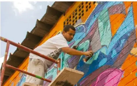 Fotografía de un artista urbano pintando un mural colorido sobre un muro, mientras trabaja en una estructura de andamio; representa la experimentación con lenguajes artísticos.