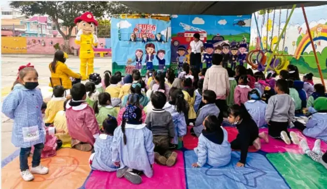 Fotografía de una función de teatro guiñol al aire libre: un títere de bombero amarillo actúa frente a un teatrino con el texto 'Bomberos al rescate' y muchos niños y niñas sentados en tapetes de colores observan. Crédito: Luis Eduardo García Medel, Tlaxcala.