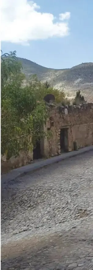 Calle empedrada en descenso con muros de piedra, monte árido y cerros al fondo bajo cielo azul.