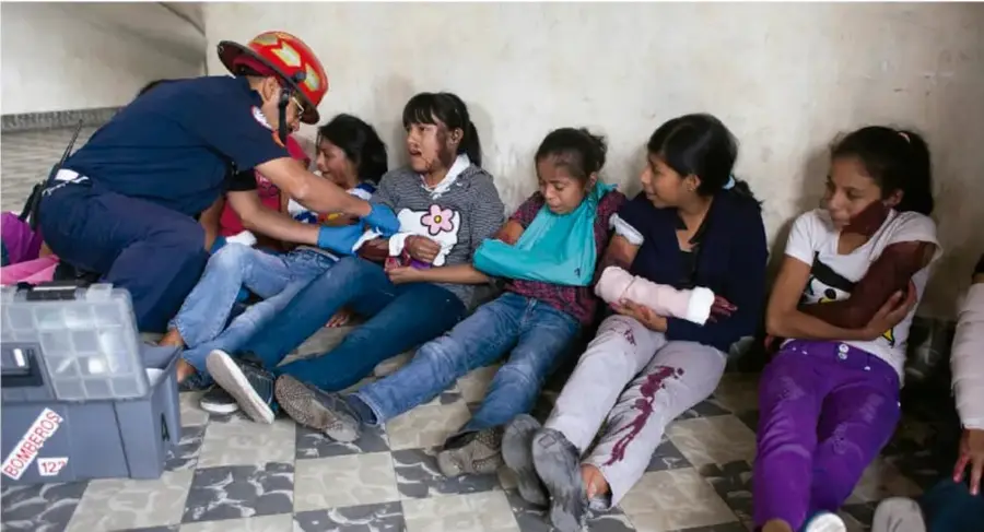 Fotografía de un simulacro de primeros auxilios: un rescatista con casco rojo y guantes atiende a varias niñas sentadas en el suelo; algunas tienen vendajes y cabestrillos. Al frente se observa un botiquín de primeros auxilios.