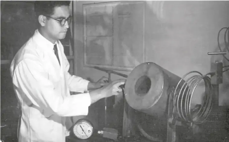 Fotografía en blanco y negro de Luis Ernesto Miramontes en el laboratorio, con bata blanca, operando un equipo de síntesis química con manómetro y bobinas de tubería.